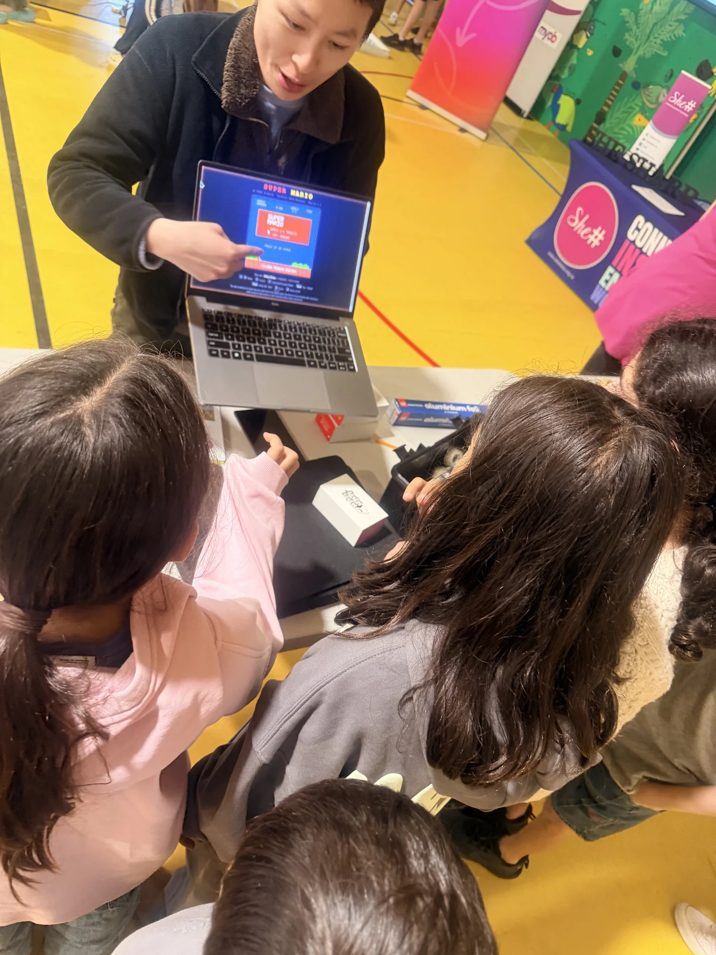 An instructor points at a laptop screen while students explore Makey Makey hardware at the workshop table